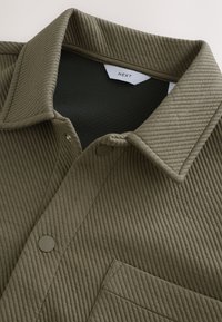 Camicia in corduroy verde oliva con colletto a punta, tasca frontale e chiusura a bottoni. Tessuto testurizzato con dettagli a righe verticali.