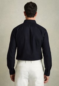 Mann mit kurzen dunklen Haaren, der ein schwarzes Langarmshirt und weiße Hosen trägt und vor einer hellgrauen Wand steht.