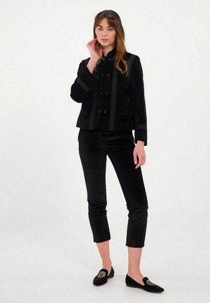 Femme debout portant une veste noire à double boutonnage, un pantalon noir court et des mocassins noirs avec un emblème doré sur fond blanc.