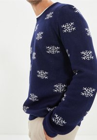 Pull bleu marine avec motifs de flocons de neige blancs, poignets et ourlet côtelés, encolure ronde et finition en maille texturée.