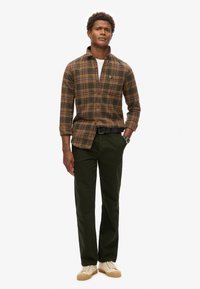 Camisa de cuadros marrón con botones y mangas largas, combinada con pantalones verdes oscuros. Zapatillas de color crema con suela texturizada. Reloj en la muñeca izquierda.