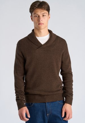 Joven con un suéter marrón de cuello chal sobre una camisa blanca y jeans azules, de pie contra un fondo claro y liso.