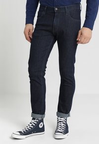 Mörkblå denimjeans med smal passform, med en klassisk femficksdesign, bältesloopar och knappstängning. Nedvikta ärmar och sneakers.