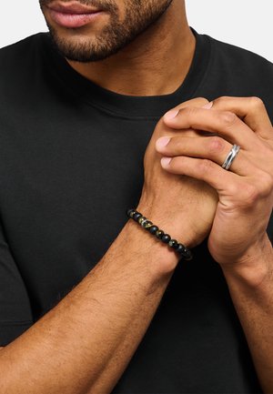 Bracciale nero con perline composto da pietre lisce opache e lucide con accenti metallici. Indossato al polso contro uno sfondo di maglietta nera.