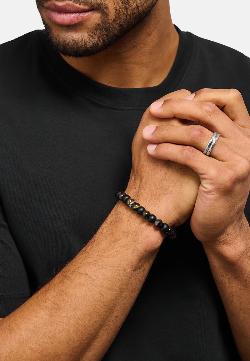 Schwarzes Perlenarmband mit glatten, matten und polierten Steinen sowie metallischen Akzenten. Getragen am Handgelenk vor schwarzem Hemdhintergrund.