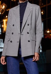 Blazer doppiopetto in bianco e nero a motivo houndstooth con grandi bottoni, rever a punta e tasche laterali, indossato sopra un dolcevita scuro.