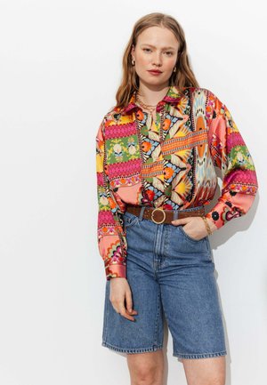 Femme portant un chemisier à motifs floraux colorés rentré dans un short en denim taille haute avec une ceinture marron, debout devant un fond blanc uni.