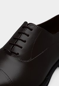 Καφέ δερμάτινο παπούτσι με λεία, γυαλιστερή επιφάνεια. Διαθέτει στρογγυλεμένη μύτη, μπροστινό δέσιμο με κορδόνια και διακριτικές ραφές κατά μήκος των ακμών.