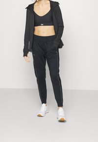 Svarta sportkläder som inkluderar en figurnära sport-bh, en lätt zip-up jacka och tapered byxor med reflexdetaljer på sidorna. Vita sneakers.