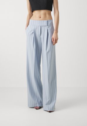 Modèle féminin portant un pantalon à pinces bleu clair à rayures fines, taille haute et jambes larges, avec des talons noirs ouverts, debout devant un fond uni.