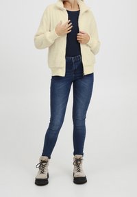 Persona con chaqueta de felpa crema con cremallera sobre una camiseta negra, jeans ajustados azul oscuro y botas beige con cordones sobre un fondo blanco.