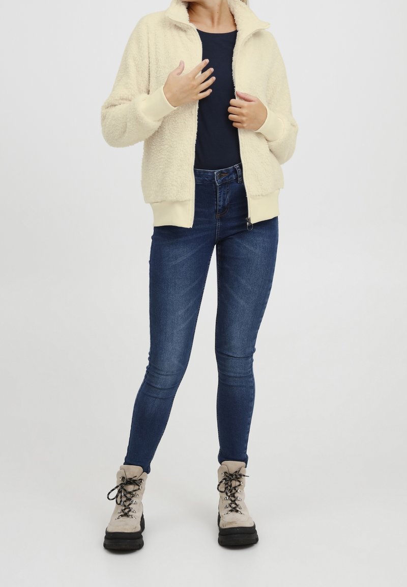Persona con chaqueta de felpa crema con cremallera sobre una camiseta negra, jeans ajustados azul oscuro y botas beige con cordones sobre un fondo blanco.