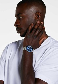 Silver bracelet watch with a blue dial and white accents, displayed on a wrist of a person wearing a plain white T-shirt.