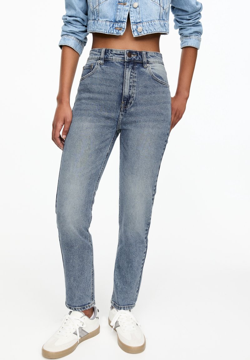 Jeans de tiro alto, de mezclilla azul claro con corte recto, que presentan bolsillos frontales y una textura desgastada, combinados con zapatillas blancas.