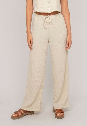 Femme portant un pantalon large beige texturé à taille coulissée et un haut court assorti à boutons, assortis à des sandales marron.