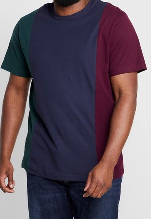 Hombre con una camiseta de manga corta con bloques de colores en verde oscuro, azul marino y granate, combinada con vaqueros azul oscuro.