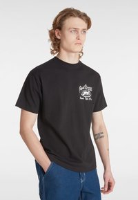 Camiseta de algodón negra con un logo gráfico blanco en el lado izquierdo del pecho. Mangas cortas y cuello redondo clásico. Combinada con vaqueros azules.