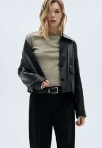 Čierna kožená skrátená bunda s detailmi na gombíkoch, nosená cez svetlozelenú košeľu. Model má tiež na sebe čierne nohavice a striebornú pracku na opasku.