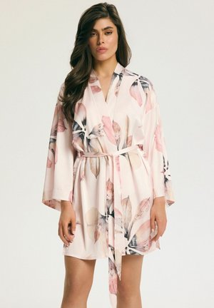 Woman wearing a light pink floral robe with wide sleeves tied at the waist, standing against a plain light background.