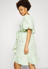 Vestido com padrão floral, com pregas em verde claro, apresentando mangas bufantes e um laço na cintura para ajuste. Tecido texturizado com barra em camadas.