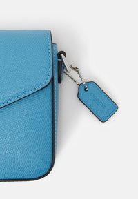Borsa in pelle blu texturizzata con chiusura a pattina, bordi neri e un piccolo tag attaccato da una catena d'argento.