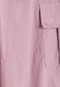 Tessuto di cotone rosa chiaro con una texture liscia; presenta una tasca sul petto e dettagli di cucitura minimal.