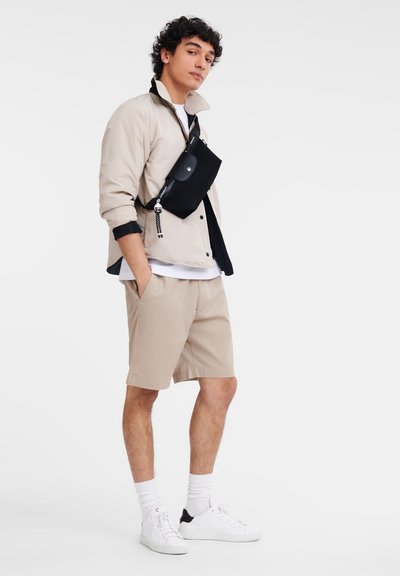Beige Shorts und eine hellbeige Jacke, die über einem weißen T-Shirt getragen wird, kombiniert mit weißen Sneakers und einer schwarzen Umhängetasche.