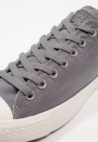 Šedá plátěná teniska s odpovídajícími tkaničkami a bílou gumovou podrážkou, detailní záběr na průchodky a šité švy.