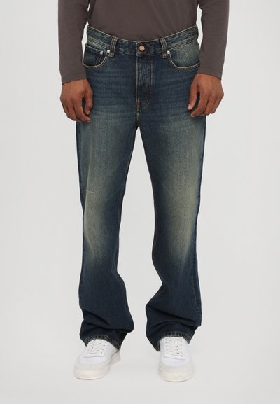 Jeans di denim blu scuro con taglio dritto, caratterizzati da una finitura sbiadita, cinque tasche e chiusura a bottone. Abbinati a sneakers bianche.