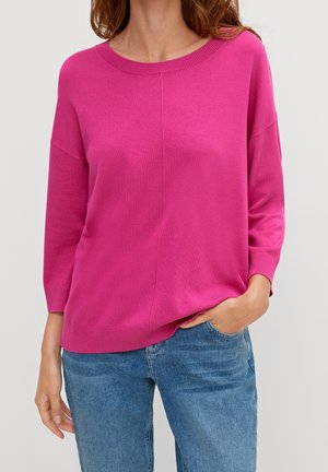 Frau trägt hellpinkes langärmeliges Pullover mit überschnittenen Schultern und blaue Jeans, eine Hand in der Tasche, vor einfachem hellem Hintergrund.