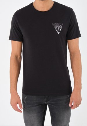 Homme portant un t-shirt noir à manches courtes avec un petit motif géométrique de montagne et de soleil sur la poitrine gauche, associé à un jean gris foncé déchiré.