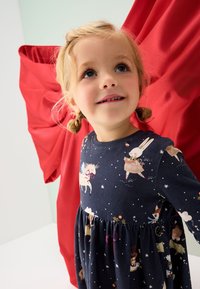 Marineblaues Kleid mit festlichen, bedruckten Tieren und Sternen, mit rundem Ausschnitt und gerafftem Rock. Gestylt vor einem roten Hintergrund.