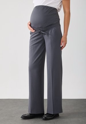 Personne enceinte portant un pantalon de maternité gris foncé avec une taille élastique et des mocassins noirs, debout sur un sol gris.