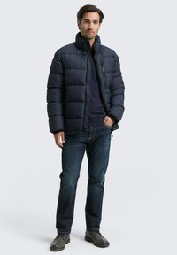 Jūras spēku pūkains cimds ar quilted dizainu, augstu apkakli un rāvējslēdzēja aizdari, kombinēts ar tumšajiem džinsiem un pelēkajiem zābakiem, stāvot uz vienkārša fona.