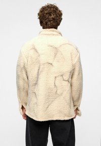 Beige fleecejas met een gestructureerd oppervlak, voorzien van een hoge kraag en een losse pasvorm. Zwarte marmerpatronen zijn overal zichtbaar.