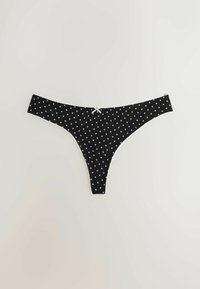 Chiloti tanga negri cu modele mici de inimi albe și o fundă albă în centrul elasticului, pe un fundal simplu și deschis la culoare.