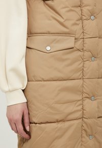 Gilet rembourré beige avec des boutons-pression argentés, doté d'une poche poitrine et d'une texture matelassée à l'avant, associé à un haut à manches longues crème.