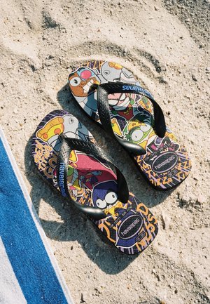 Een paar kleurrijke Havaianas slippers met cartoonfiguren op een zandstrand naast een blauw-wit gestreepte handdoek.