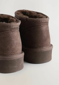 Braune Wildleder-Stiefeletten mit einem weichen Fellfutter, runder Form und flacher Gummisohle. Die sichtbaren Nahtdetails an den Seiten sind auffällig.