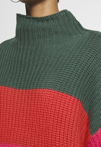 Neulepusero, jossa on korkea kaulus ja vihreitä, punaisia ja vaaleanpunaisia vaakaraitoja. Teksturoitu kangas, jossa näkyvät ompeluyksityiskohdat.