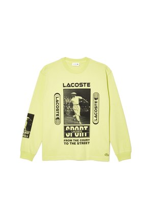 Chemise à manches longues vert clair avec logos Lacoste noirs, un graphique vintage de joueur de tennis et le texte "SPORT de la cour à la rue."