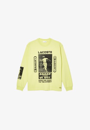 Chemise à manches longues vert clair avec logos Lacoste noirs, un graphique vintage de joueur de tennis et le texte "SPORT de la cour à la rue."