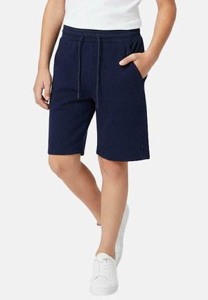 Persoon draagt marineblauwe knielange short met trekkoord in de taille, witte sneakers en een wit shirt, staand met één hand in de zak.