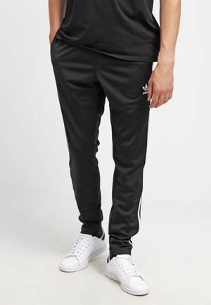 Personne portant un pantalon de survêtement noir Adidas avec des bandes blanches et un logo, associé à des baskets blanches, debout devant un fond uni.