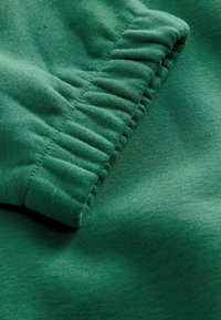 Un material textil verde cu o textură moale, având detalii pliate de-a lungul marginii. Materialul pare confortabil și este țesut strâns.