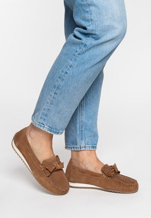 Voeten met lichtblauwe spijkerbroek en bruine suède mocassins met strikdetail en witte zolen tegen een witte achtergrond.