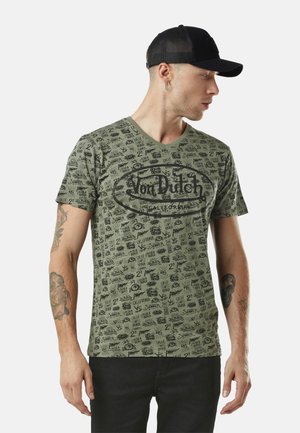 Homme portant un t-shirt graphique Von Dutch olive, une casquette noire en maille et un pantalon noir, regardant vers sa droite avec des tatouages visibles sur les deux bras.