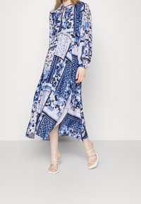 Midi-jurk met blauw patroon, met bloemen- en ruitmotieven, lange mouwen en een striksluiting bij de hals. Gecombineerd met transparante sandalen met bandjes.