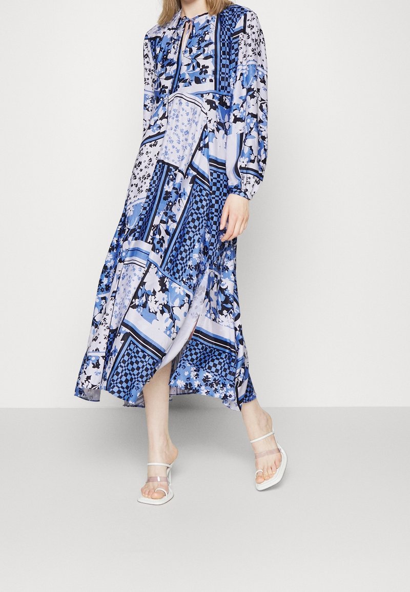 Midi-jurk met blauw patroon, met bloemen- en ruitmotieven, lange mouwen en een striksluiting bij de hals. Gecombineerd met transparante sandalen met bandjes.