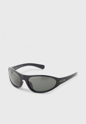 Gafas de sol envolventes negras con lentes oscuras y un pequeño logo de metal de la marca en la patilla sobre un fondo blanco.